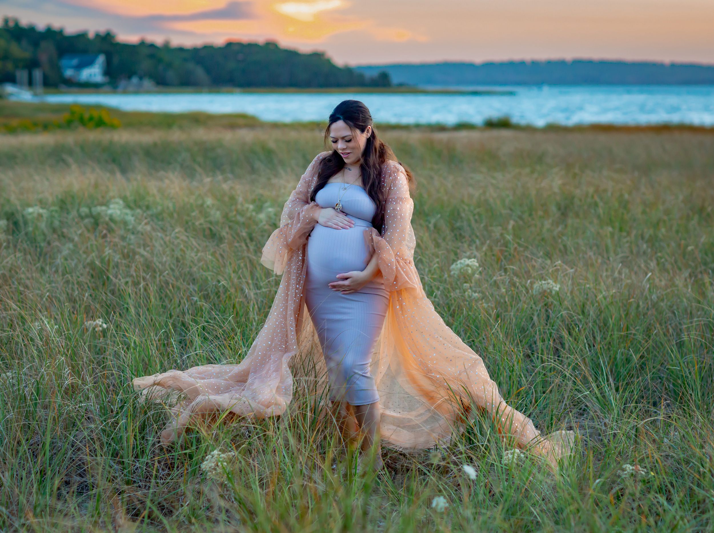 Pregnant woman in gray dress and peach robe poses in golden field by water at sunset.