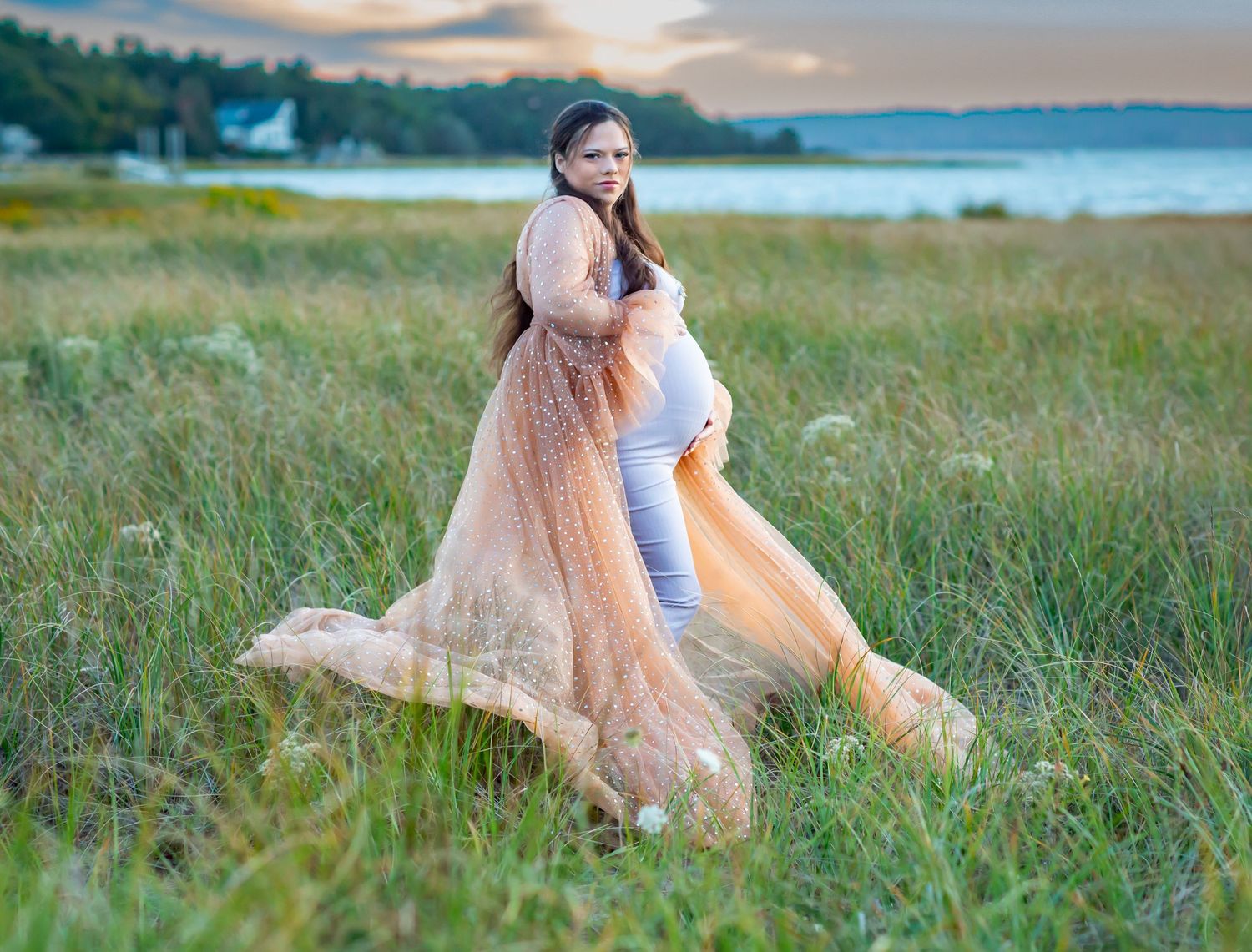 Pregnant woman in flowing peach gown poses in green meadow near water at sunset.
