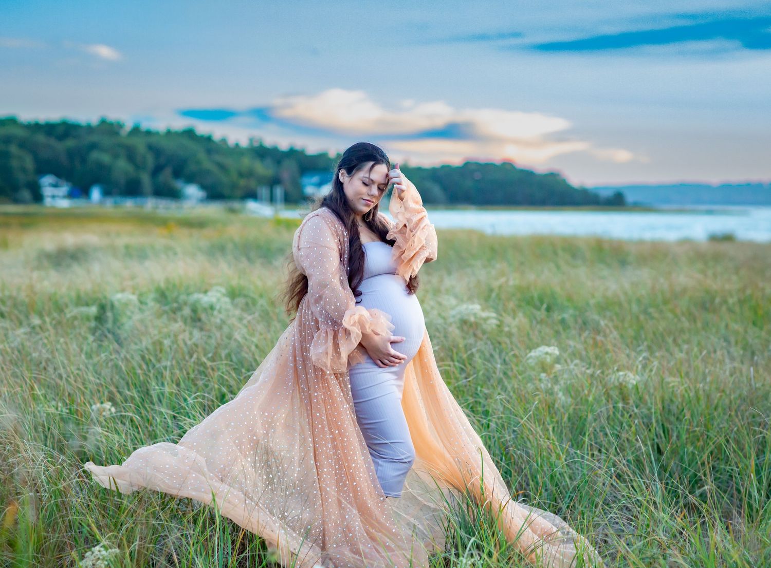 Pregnant woman in white dress with partner in peach gown embracing in a scenic grassy field near water.