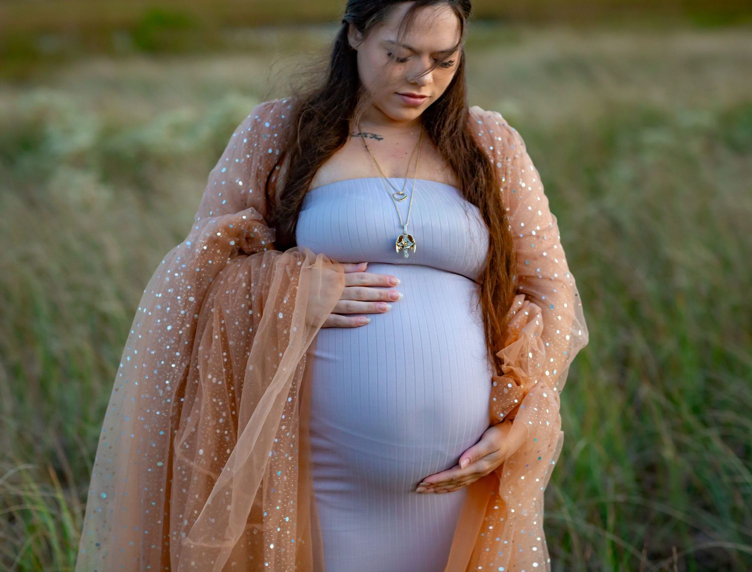 Pregnant woman in gray dress and peach tulle robe cradling baby bump in a field.