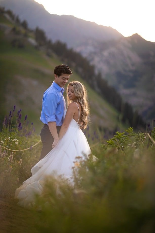 Couple embracing in a mountain meadow at golden hour, bride in white gown, wildflowers and peaks in background.