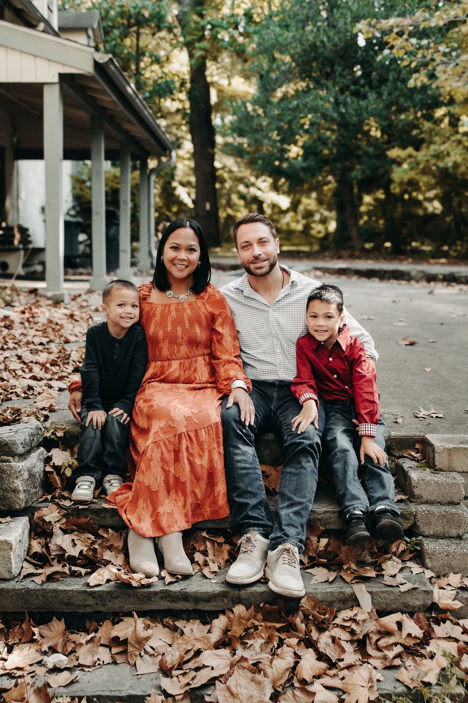 Family of four smiling on stone steps surrounded by autumn leaves near a wooded home in fall.