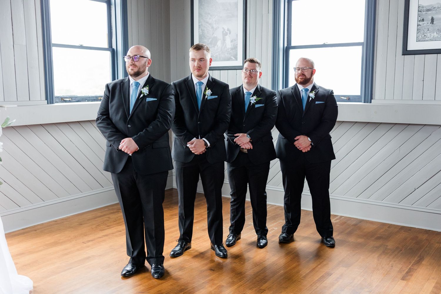 Four groomsmen in black suits with light blue ties standing together indoors before a wedding ceremony.