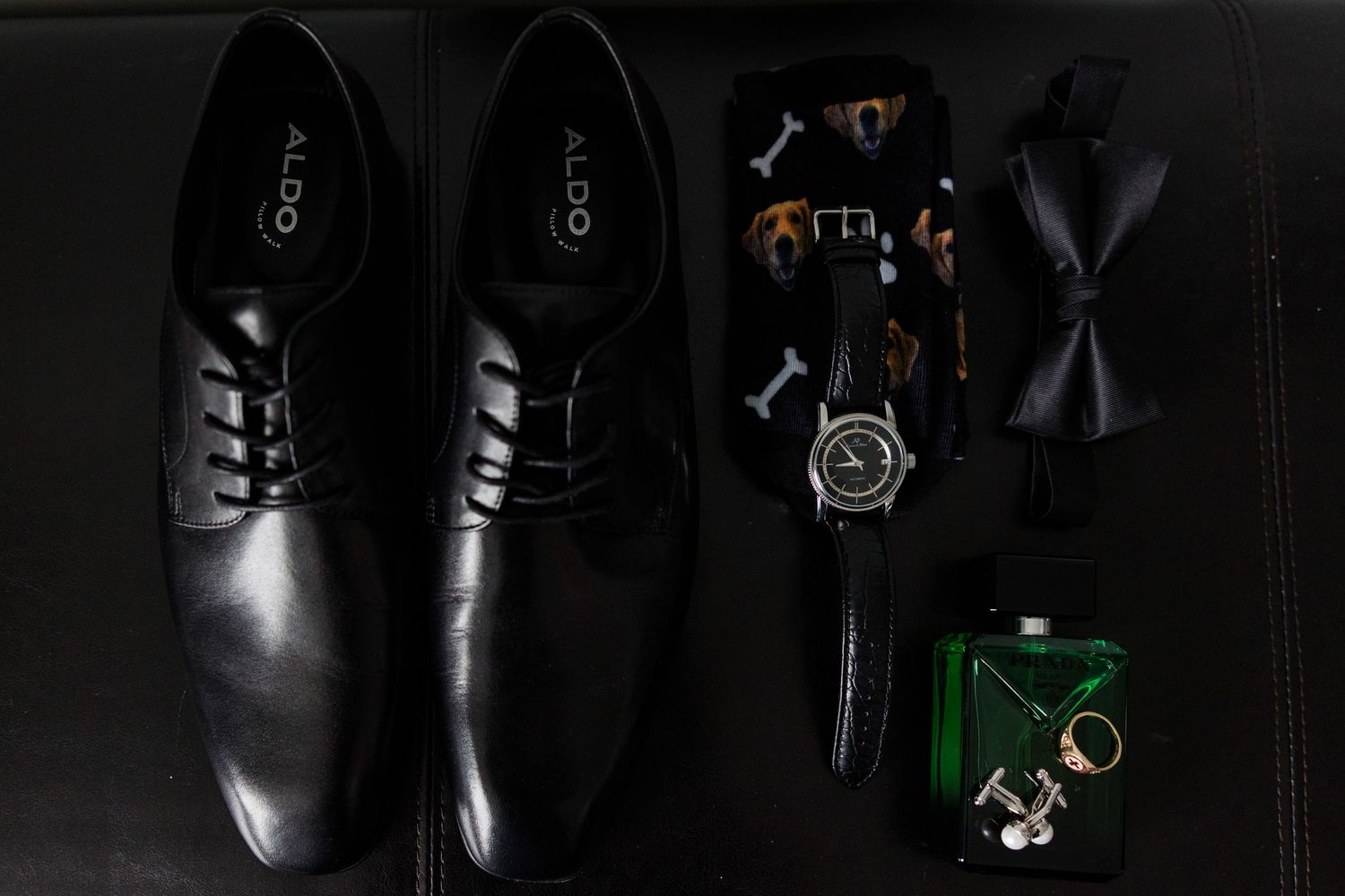 Black ALDO dress shoes displayed with a watch, bow tie, cufflinks, and cologne on a dark surface.