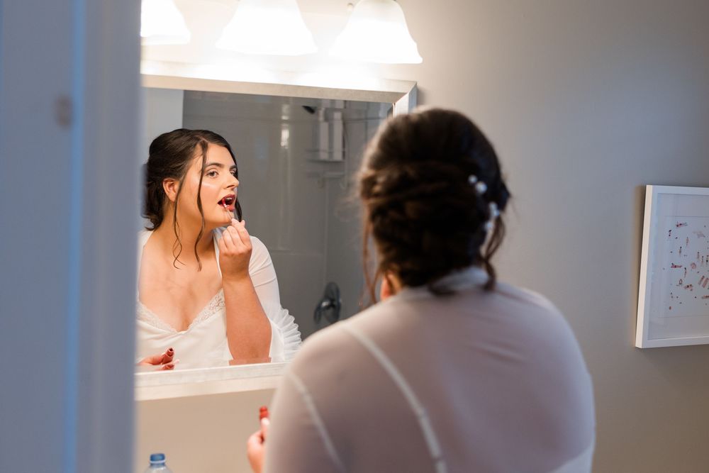 Bride applying red lipstick while looking in a bathroom mirror during wedding day preparations.
