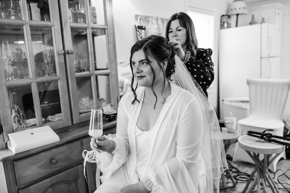 Bride in white robe holds champagne while hairstylist works on her updo in a cozy room, black and white photo.