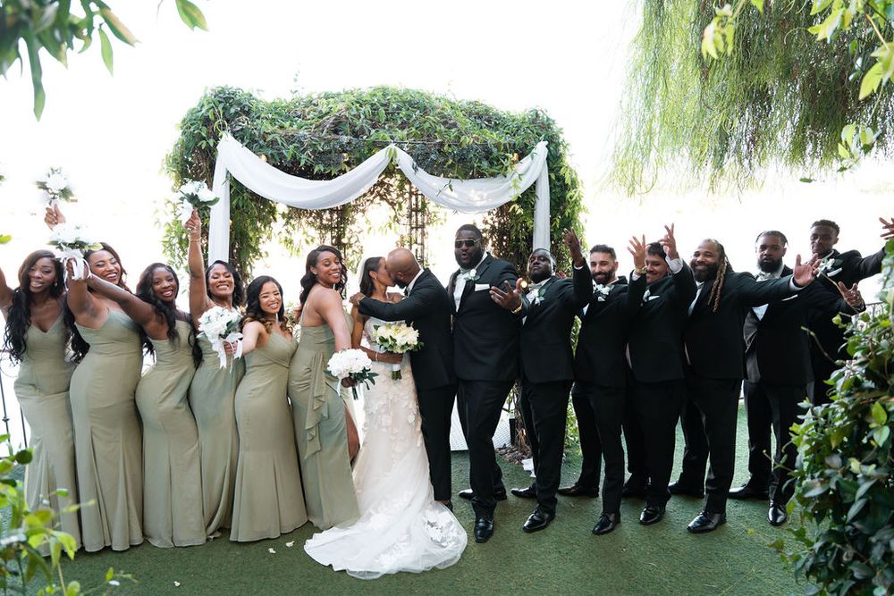 Wedding party celebrates outdoors under floral arch, bridesmaids in sage gowns and groomsmen in black tuxedos cheering.