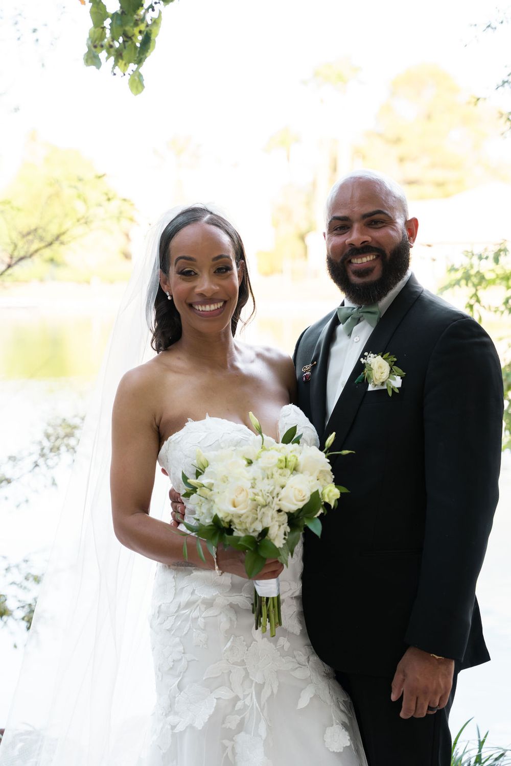 Bride and groom smiling outdoors, bride in strapless lace gown holding white bouquet, groom in black tuxedo with green bow tie.