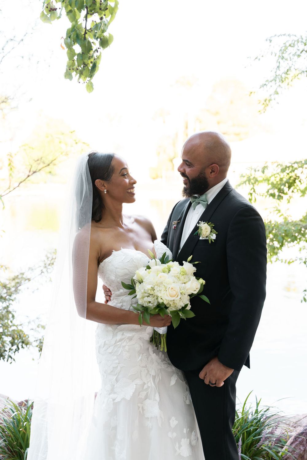 Bride and groom share a loving gaze outdoors, she holds white roses bouquet, wearing strapless gown with veil, he in black tuxedo.