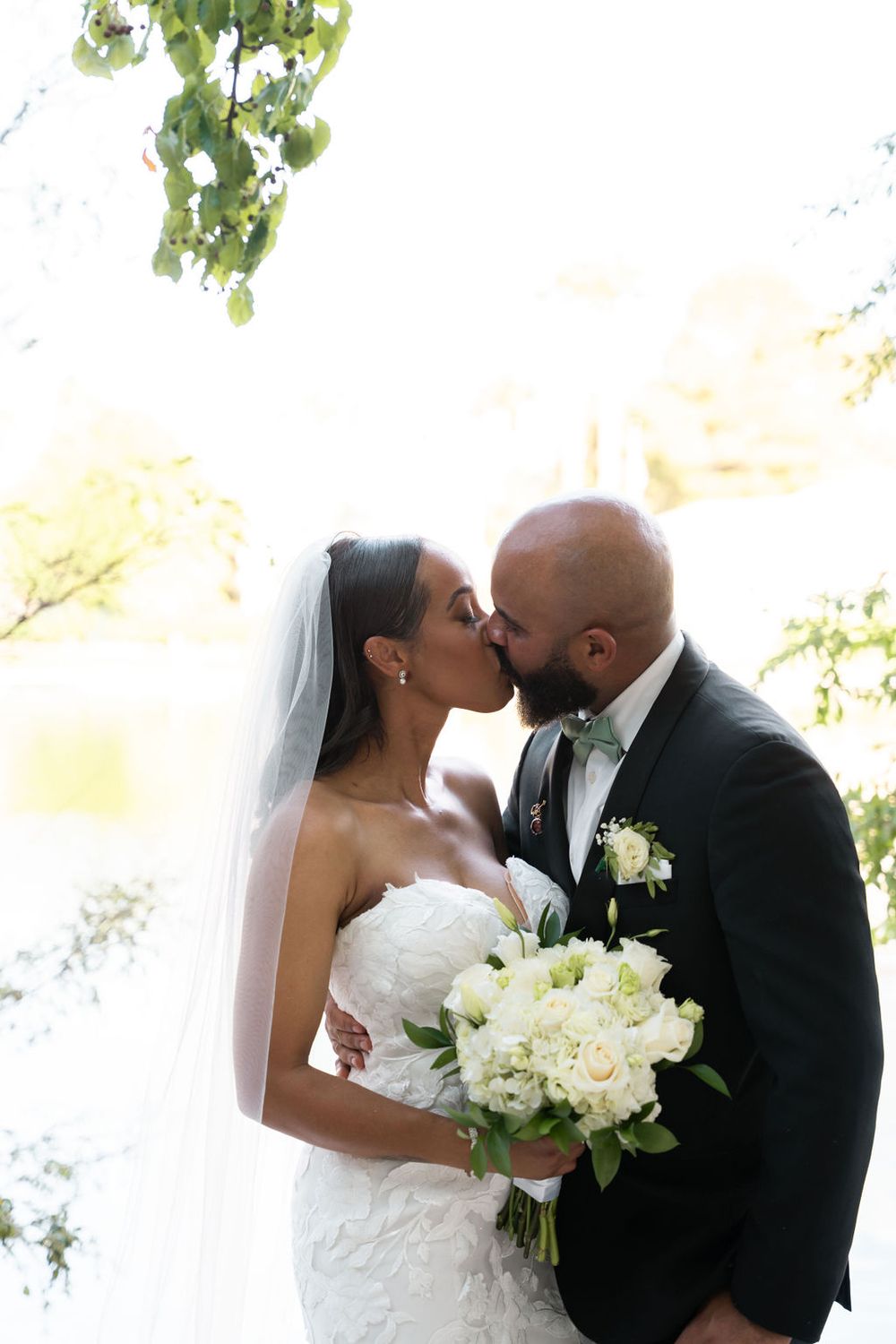 Bride and groom share a romantic kiss outdoors, bride holding white floral bouquet in elegant strapless lace gown with veil.