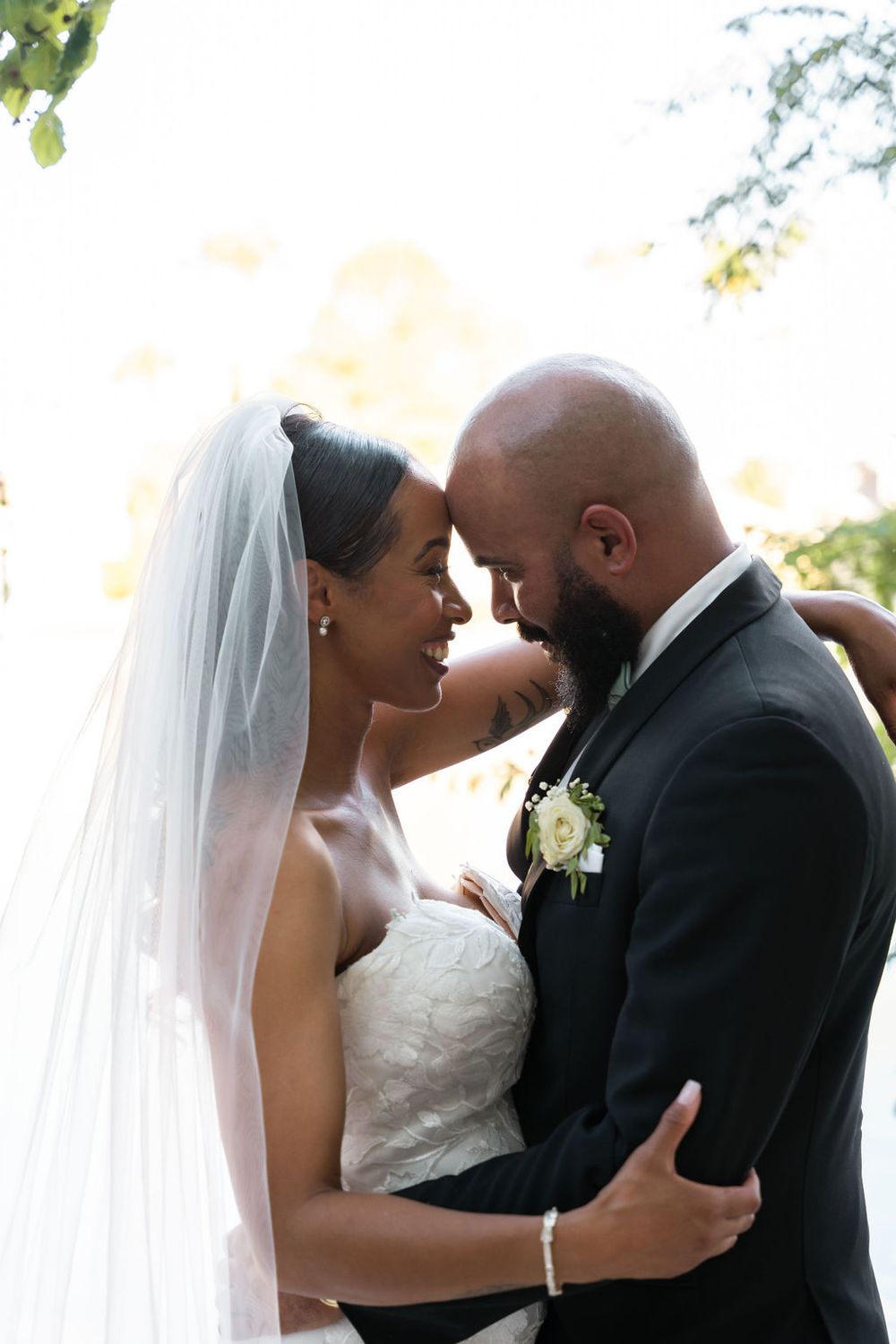 A bride and groom share an intimate forehead touch outdoors, she in a white gown with veil, he in a dark suit with boutonniere.