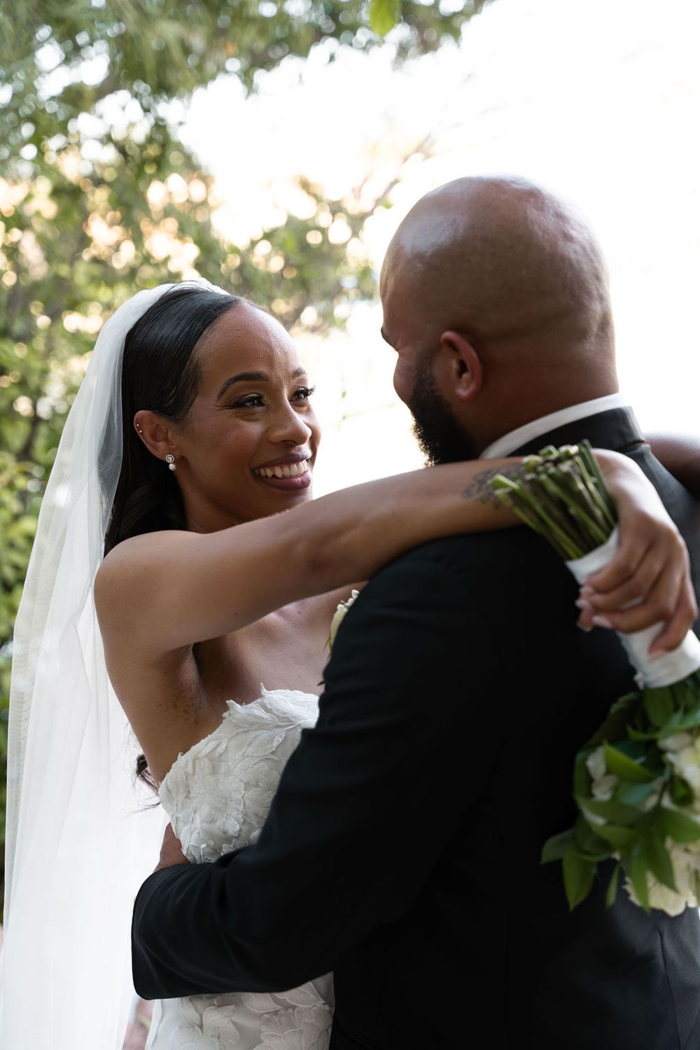 A joyful bride in a white gown and veil embraces her groom in a black suit outdoors at their wedding.