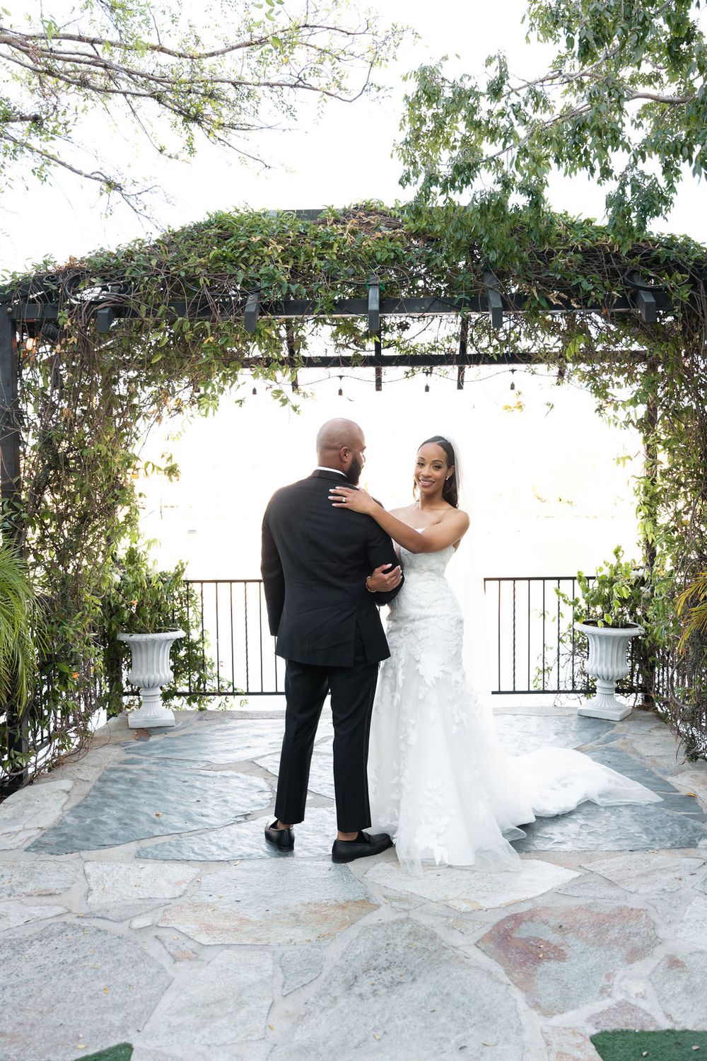 A bride and groom share their first dance under a lush vine-covered pergola on a stone patio at an outdoor wedding.
