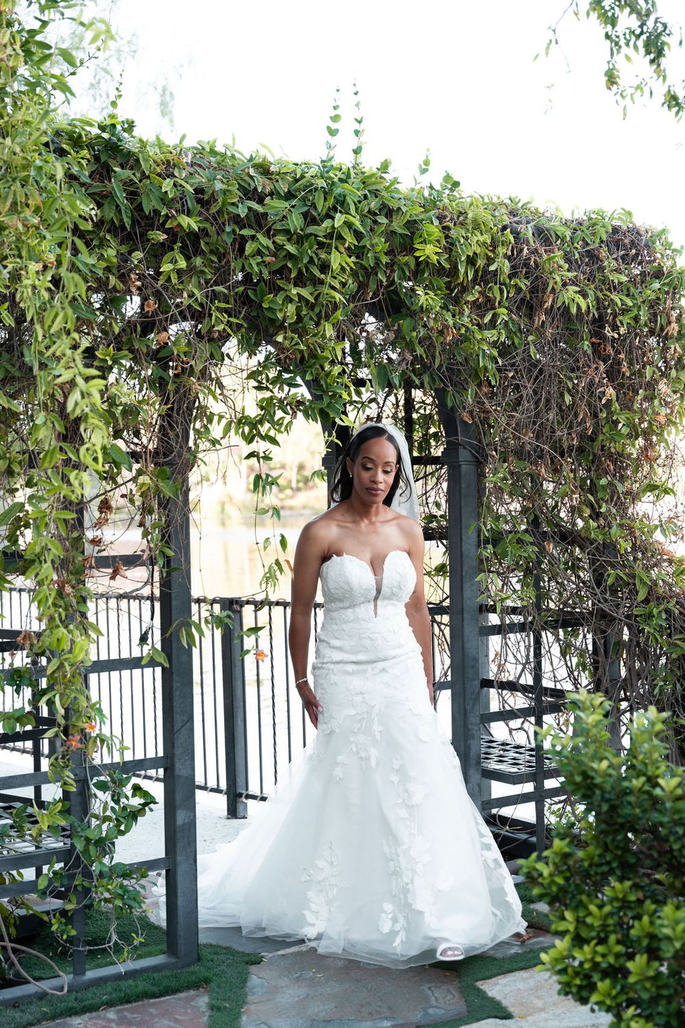 Bride in strapless lace mermaid wedding gown standing under ivy-covered pergola arch in outdoor garden setting.