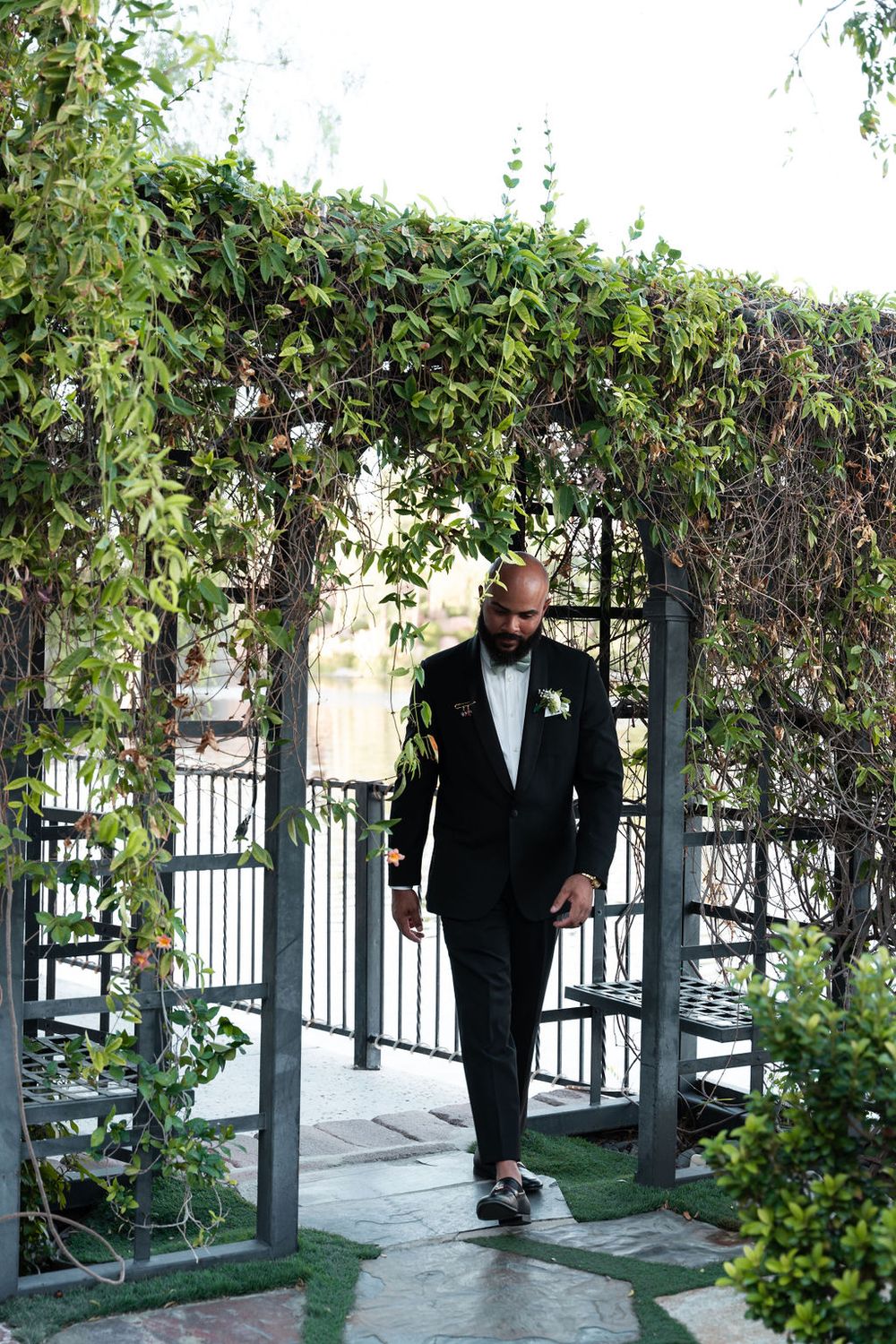 Groom in black tuxedo with boutonniere walks through ivy-covered garden arch at outdoor wedding venue.