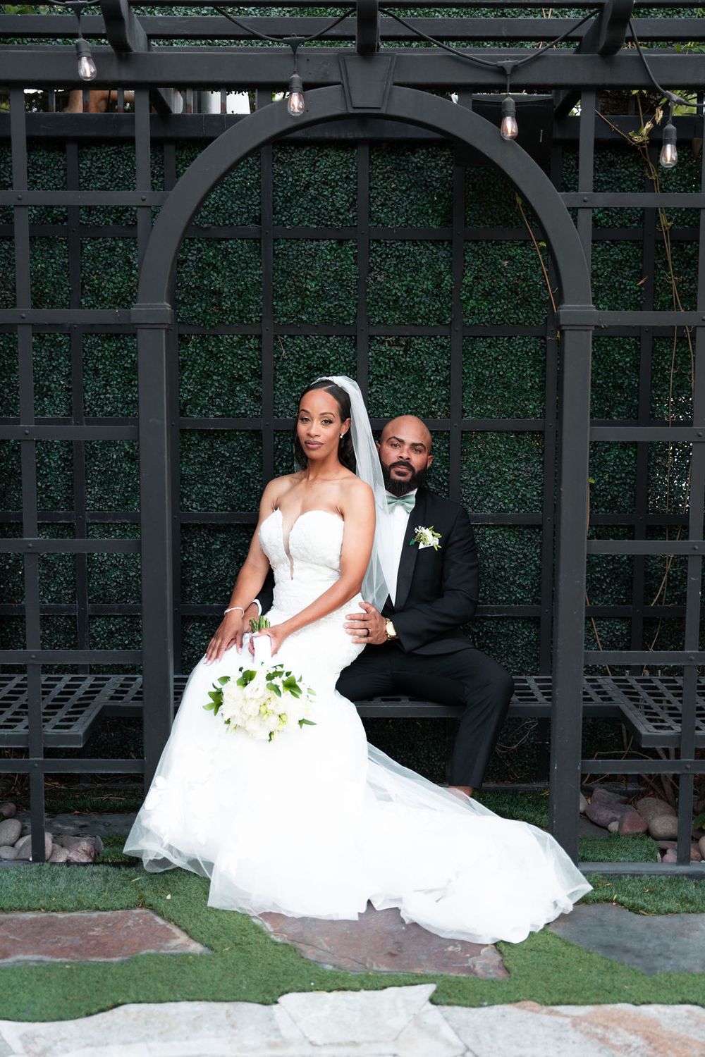 Bride in white strapless gown and groom in black tuxedo pose under a dark arch with green hedge backdrop.