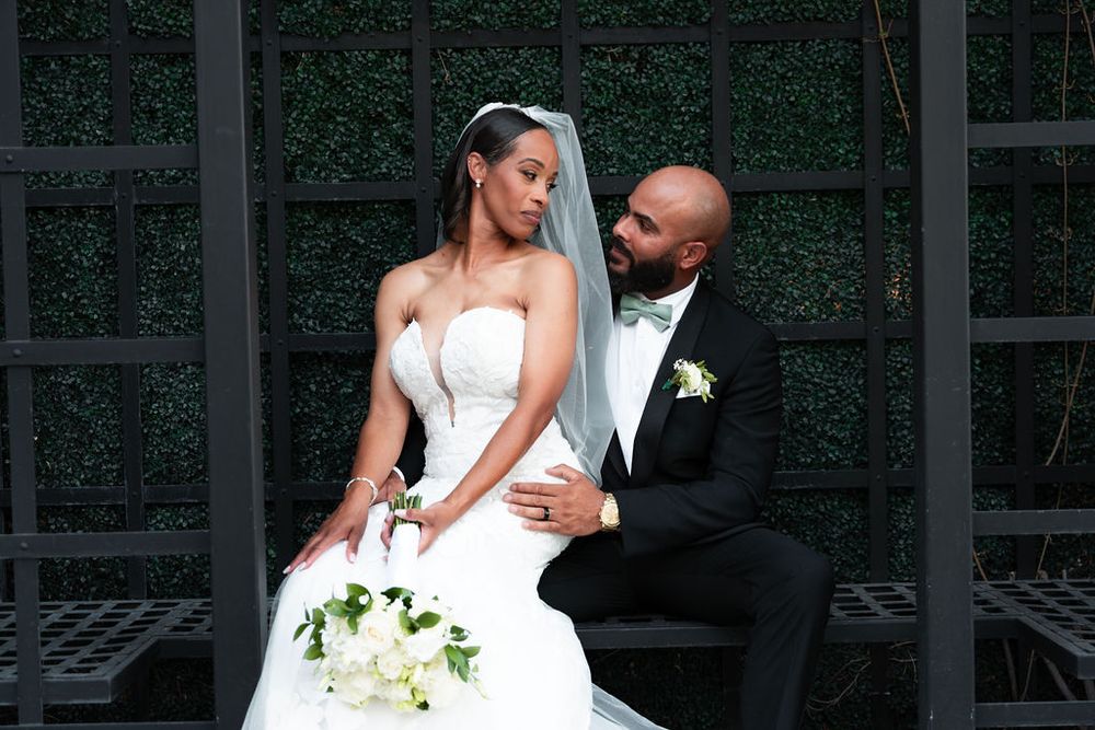 Bride in white strapless gown with veil and groom in black tuxedo pose together at an outdoor wedding.