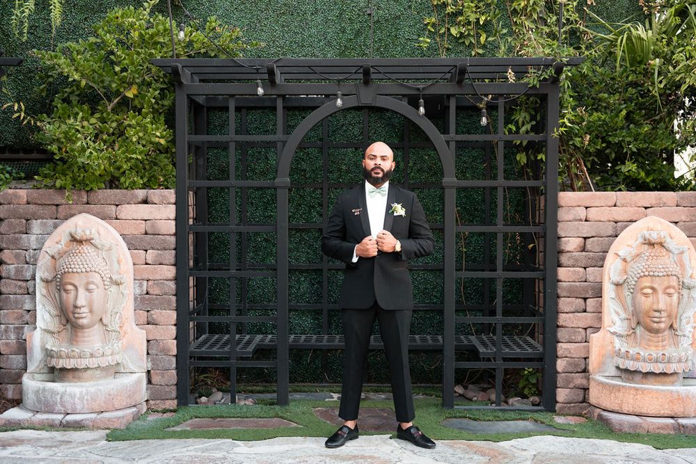Groom in black tuxedo stands confidently under a dark wooden arch flanked by stone Buddha sculptures.