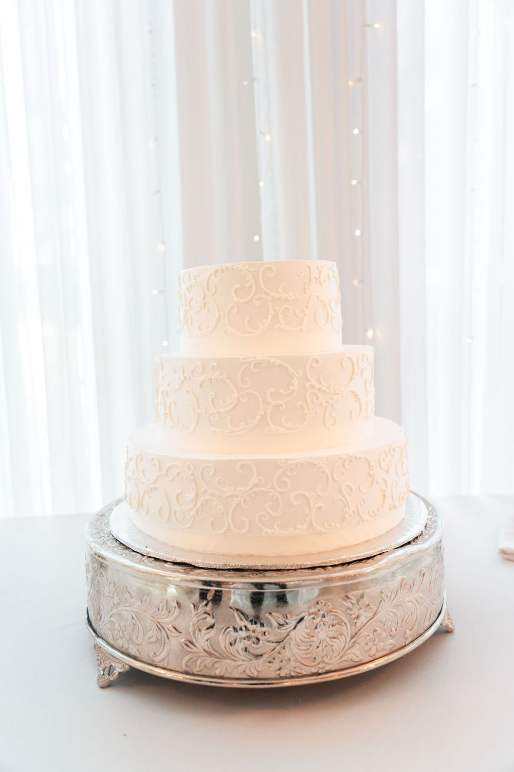 Elegant four-tier white wedding cake with scroll designs on ornate silver cake stand.