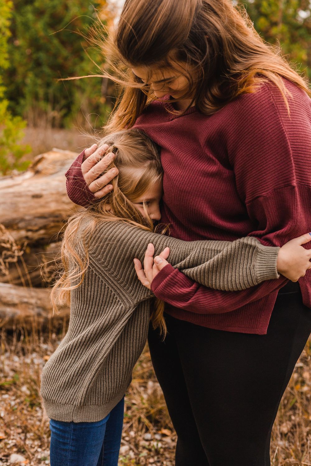 A mother in a burgundy sweater tenderly embraces her young daughter outdoors in an autumn setting.