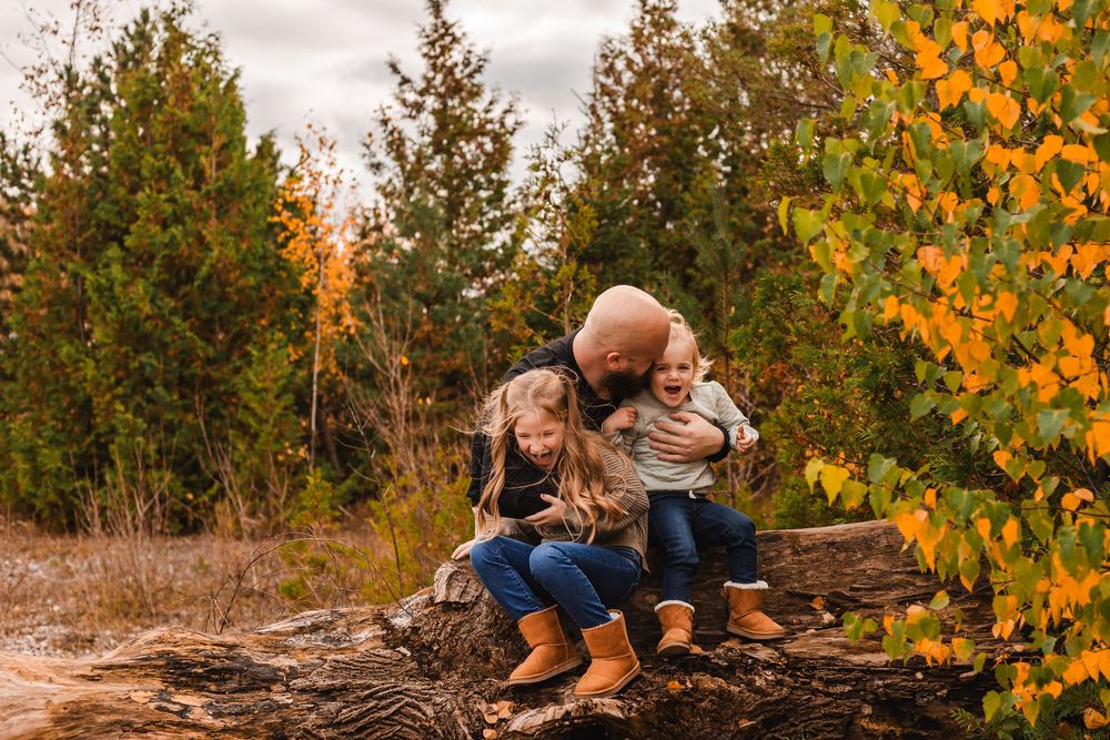 Parent with two young children sitting on a log in an autumn forest with colorful yellow and green foliage.