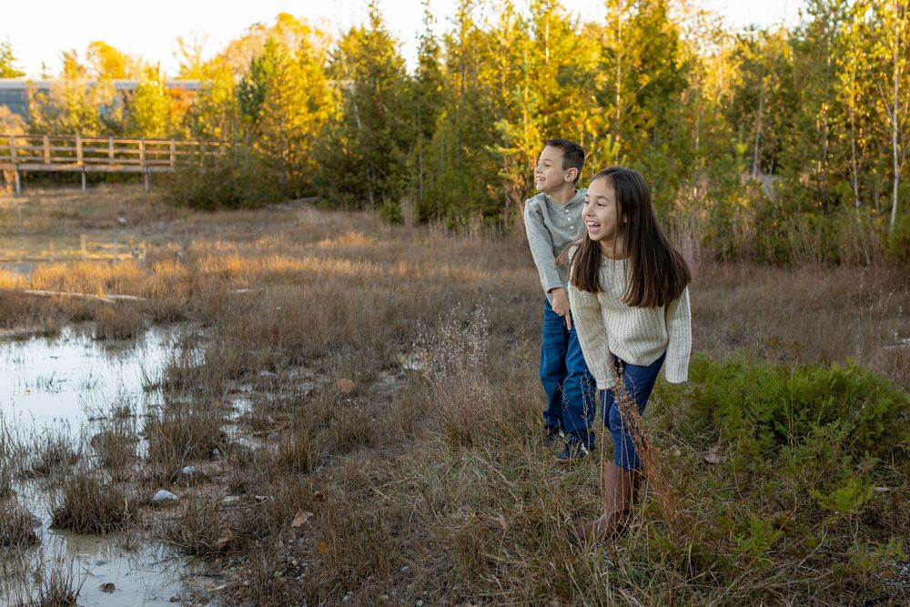 Couple embracing outdoors in a golden autumn meadow near a pond, surrounded by tall grasses and trees at sunset.