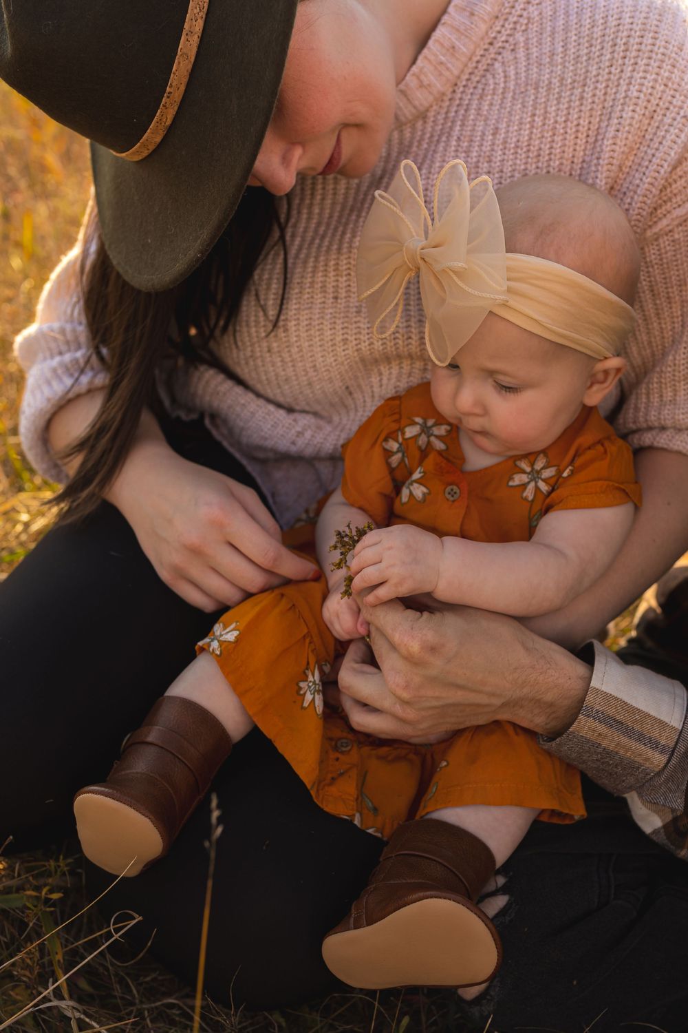 A family with a baby girl in an orange floral dress and bow headband sits outdoors in warm autumn light.