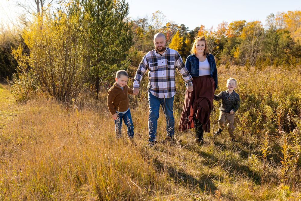 Family of four walks through golden autumn field, parents holding children's hands with colorful fall foliage in background.