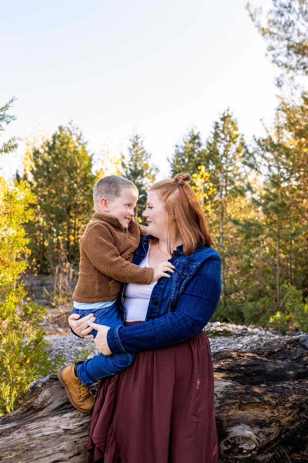 A mother with red hair holds her laughing young son outdoors among autumn trees in a warm family portrait.