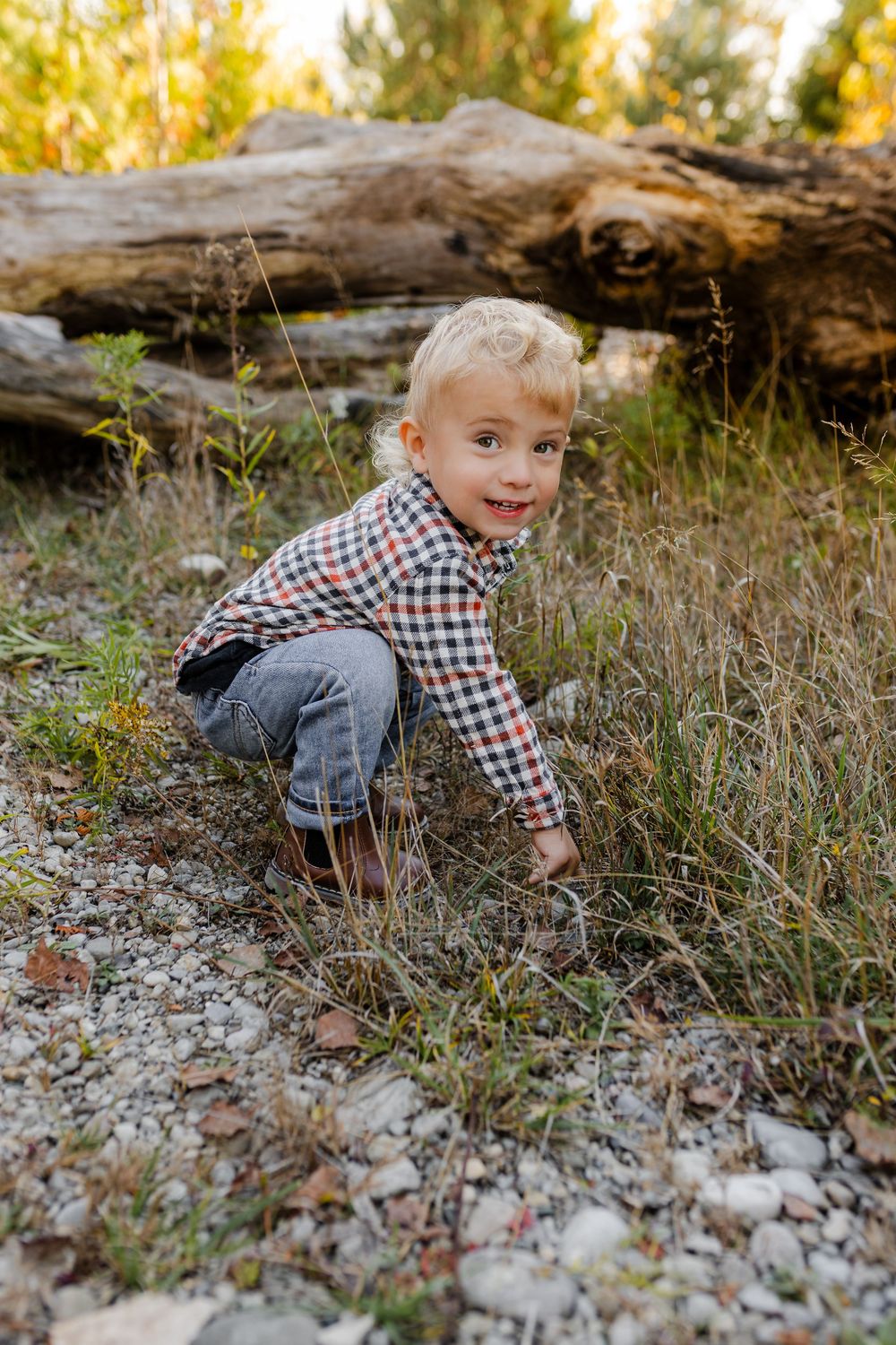 Blonde toddler boy in plaid shirt crouching on rocky ground in a natural outdoor setting with fallen logs.