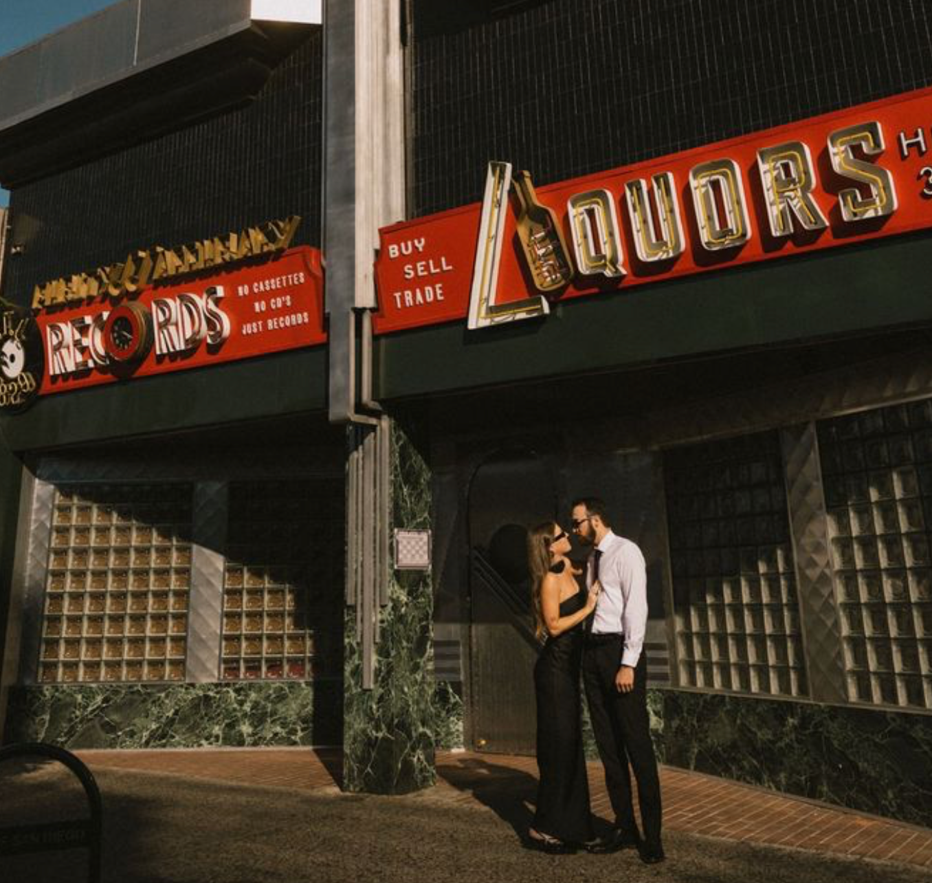 Couple embracing outside a vintage liquors store with retro neon signage in an urban setting.