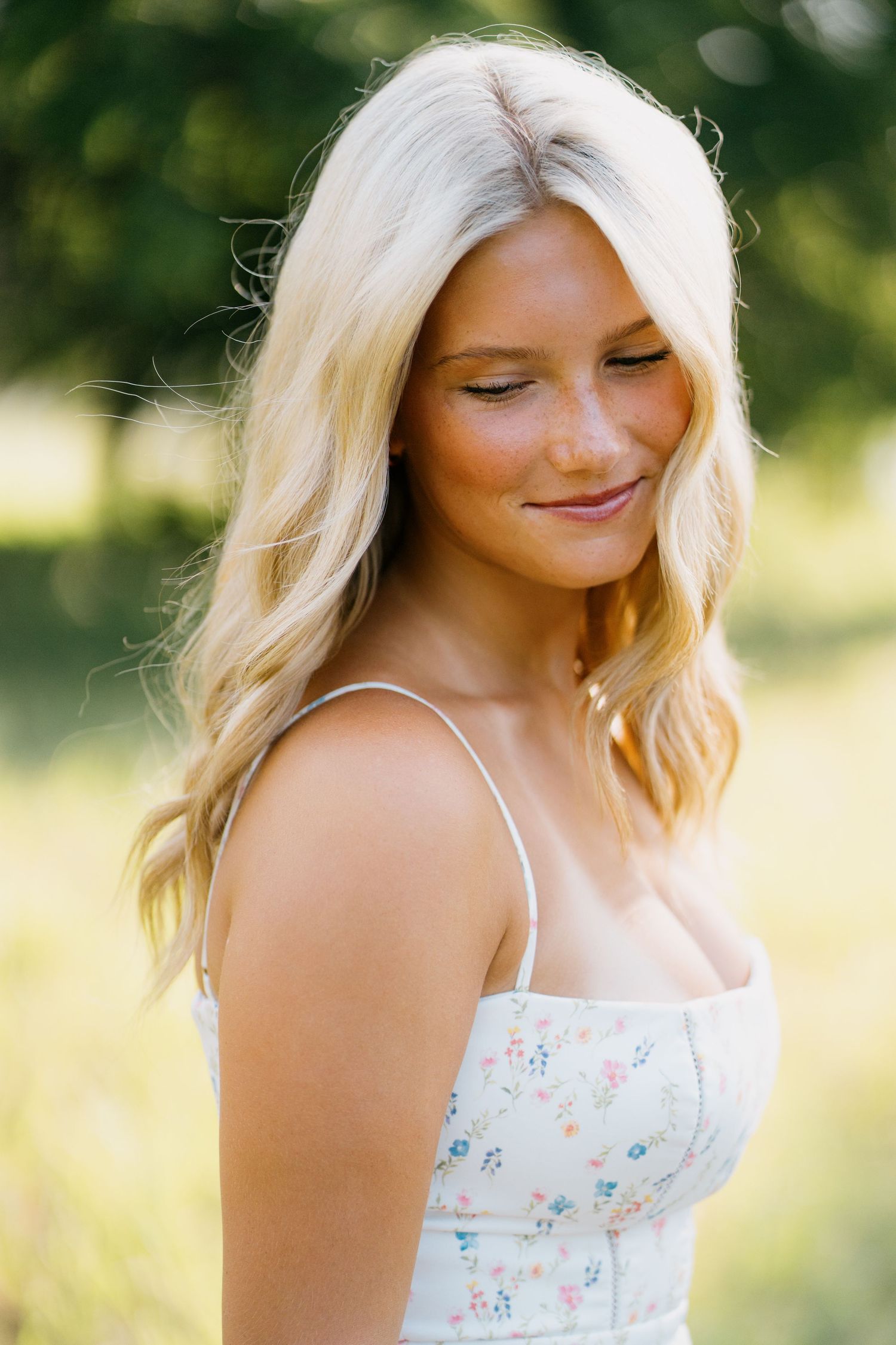 Smiling blonde woman with freckles wearing a floral sundress outdoors in a sunny green garden setting.