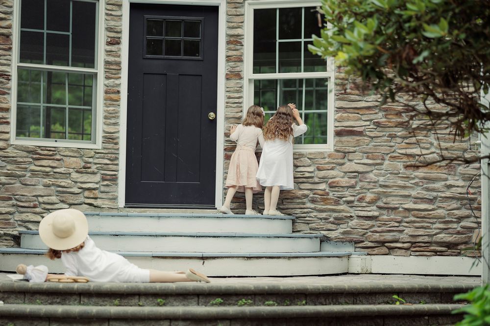 Two children in white dresses stand at a stone house door while a woman in a hat relaxes on the steps.