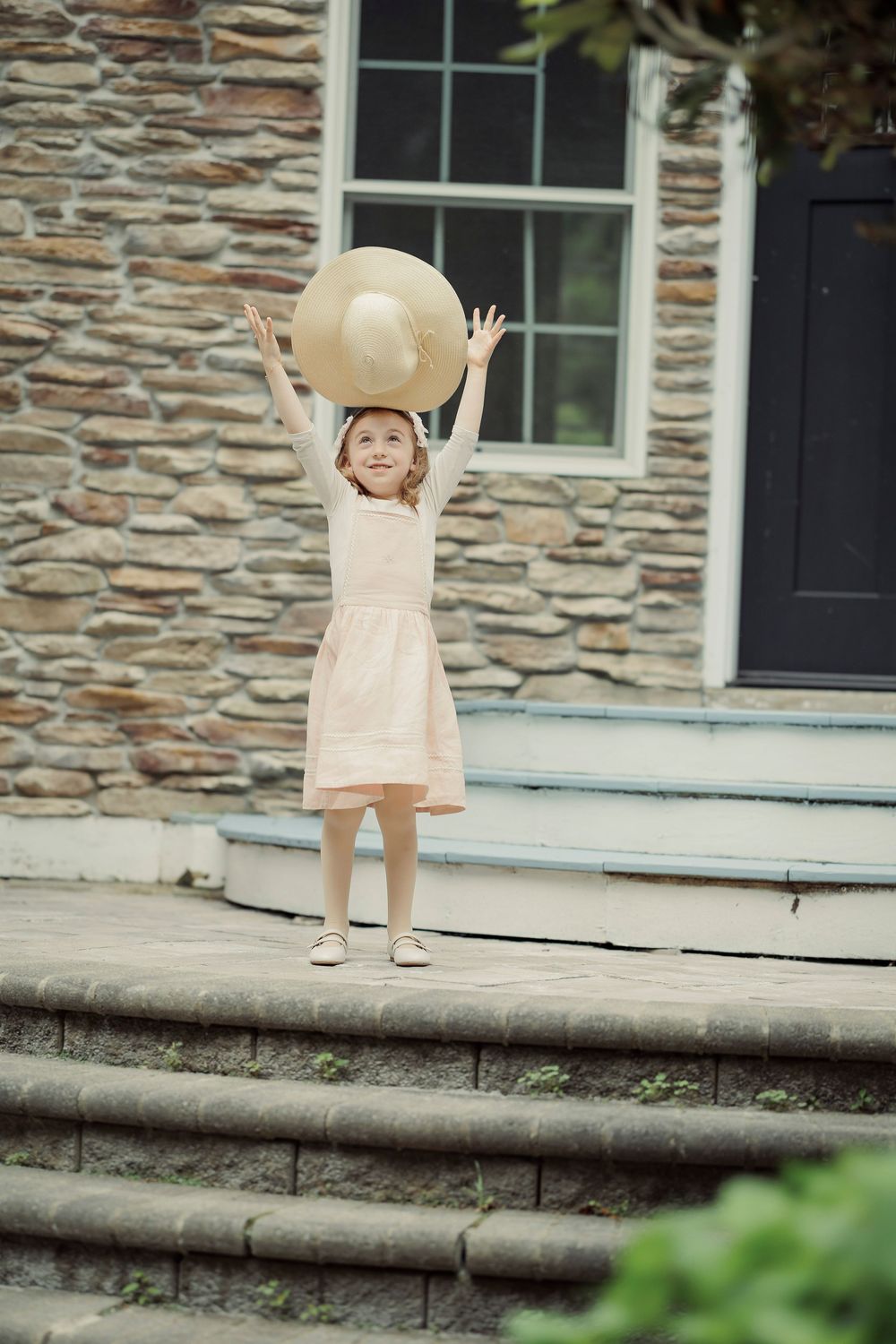 Young girl in pink dress raising straw hat above her head on stone steps outside a historic building.