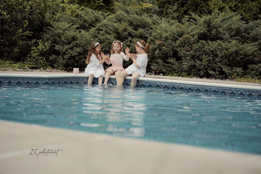 Three girls in white dresses sit poolside, laughing together with lush green trees in the background.