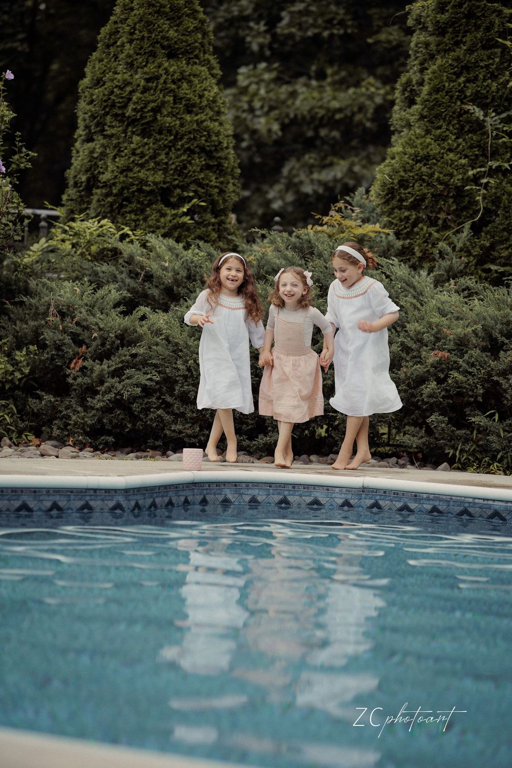 Three young girls in white and pink dresses laugh and jump together beside a pool surrounded by lush green trees.