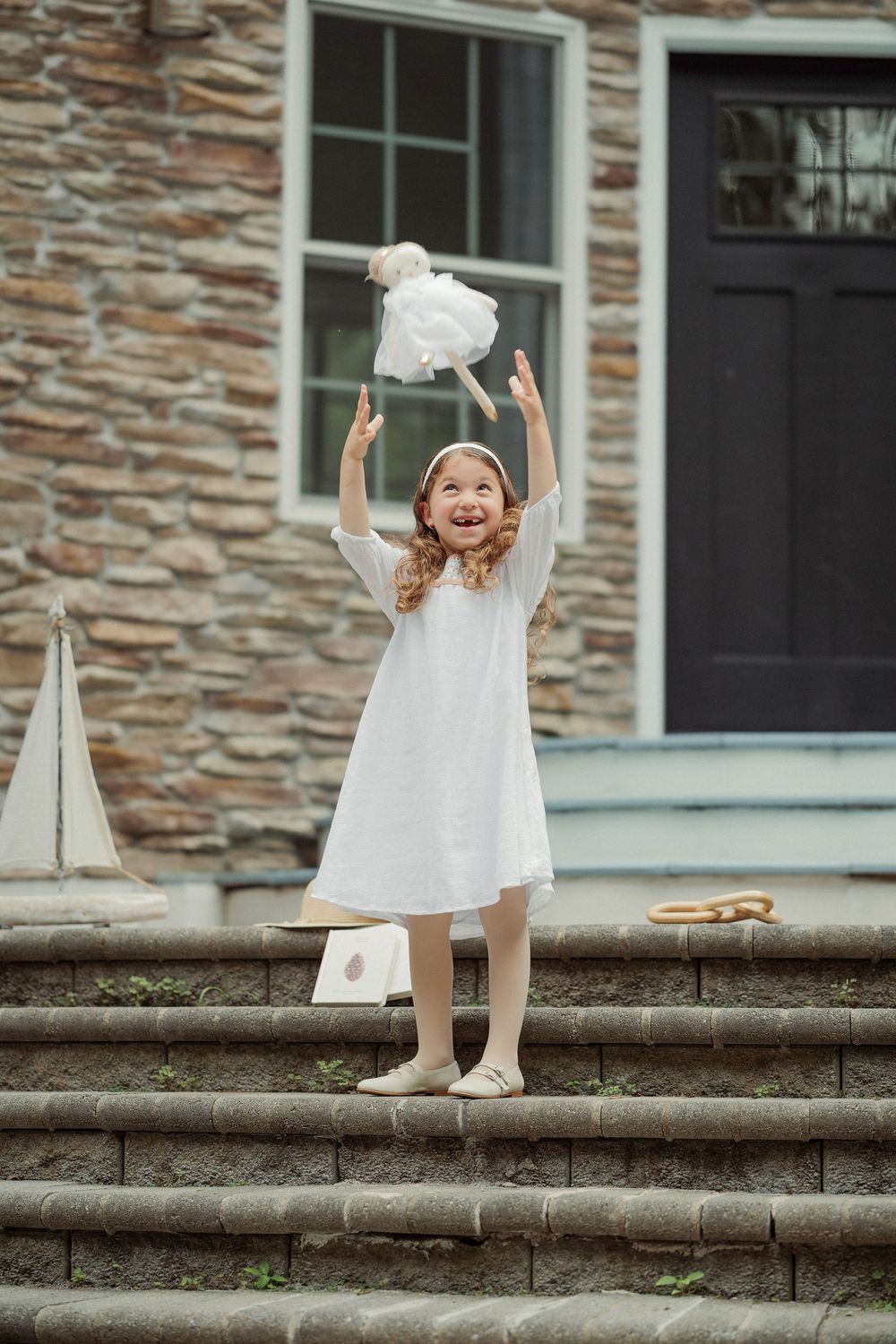 Young girl in white dress joyfully tossing a flower on stone steps outside a historic building.