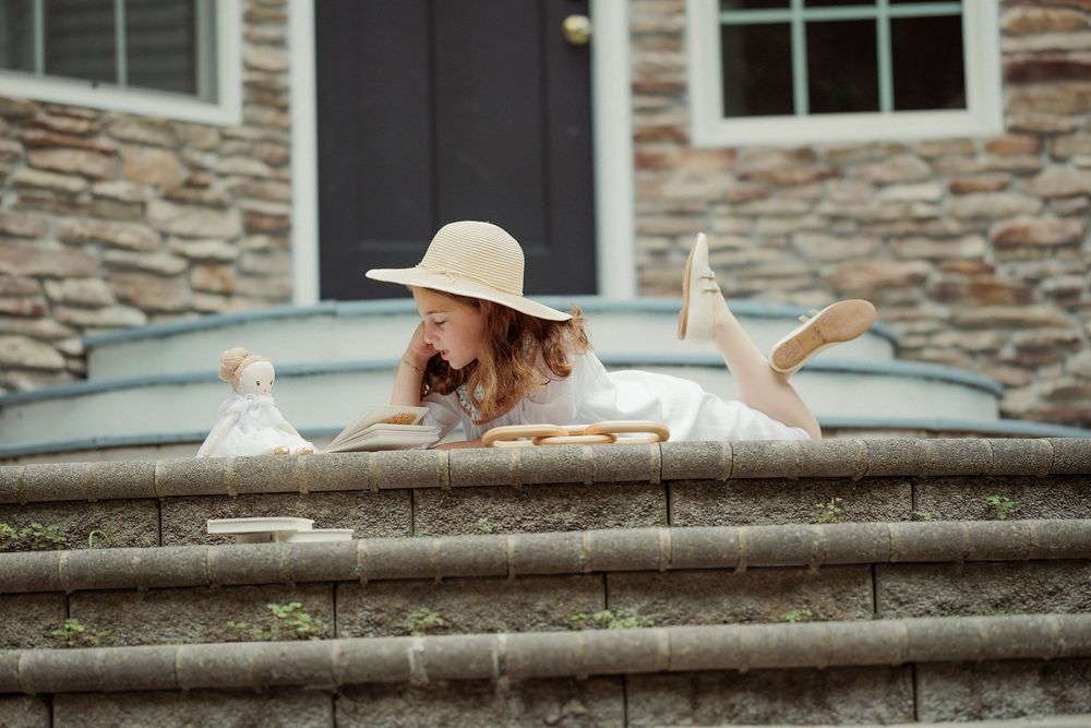 Woman in white dress and sun hat reading a book while lying on stone steps outside a building.