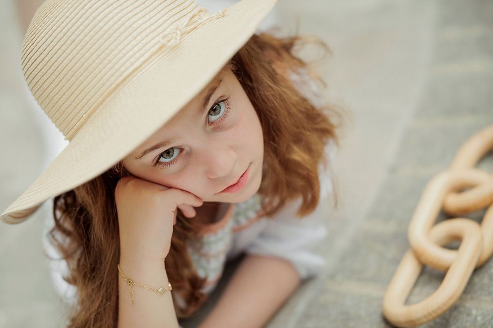 A young girl with curly hair wearing a wide-brim straw hat rests her chin on her hand, gazing upward outdoors.