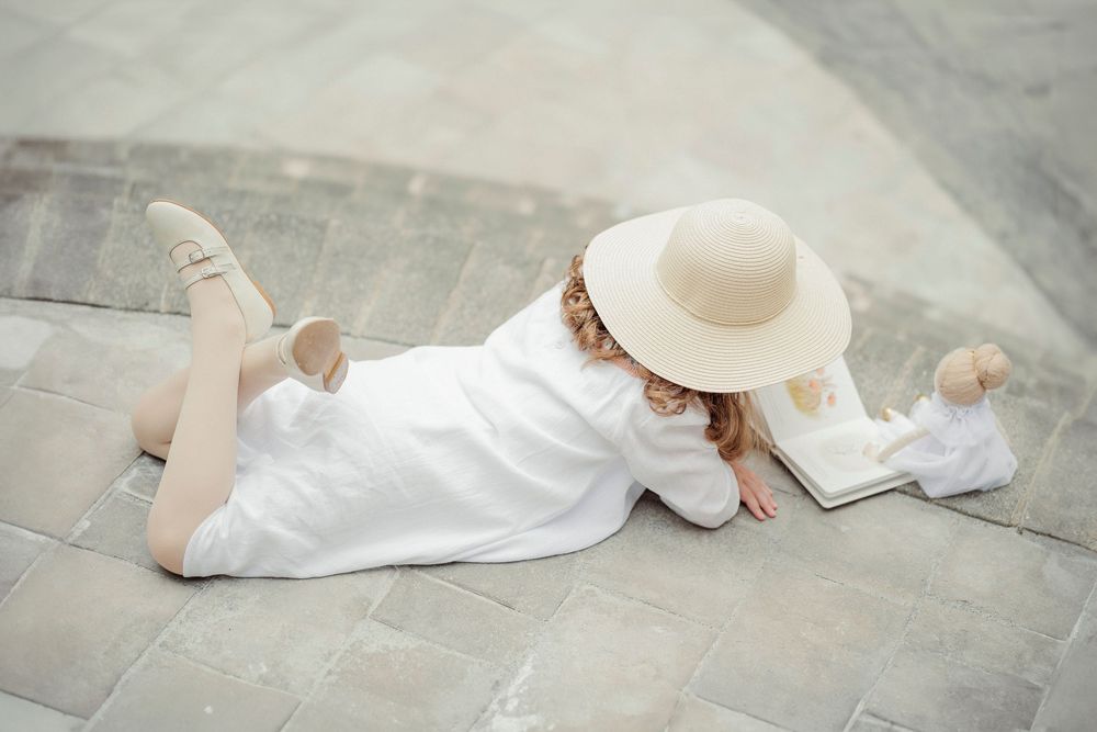 A young girl in a white dress and sun hat lies on stone steps, reading a book outdoors.