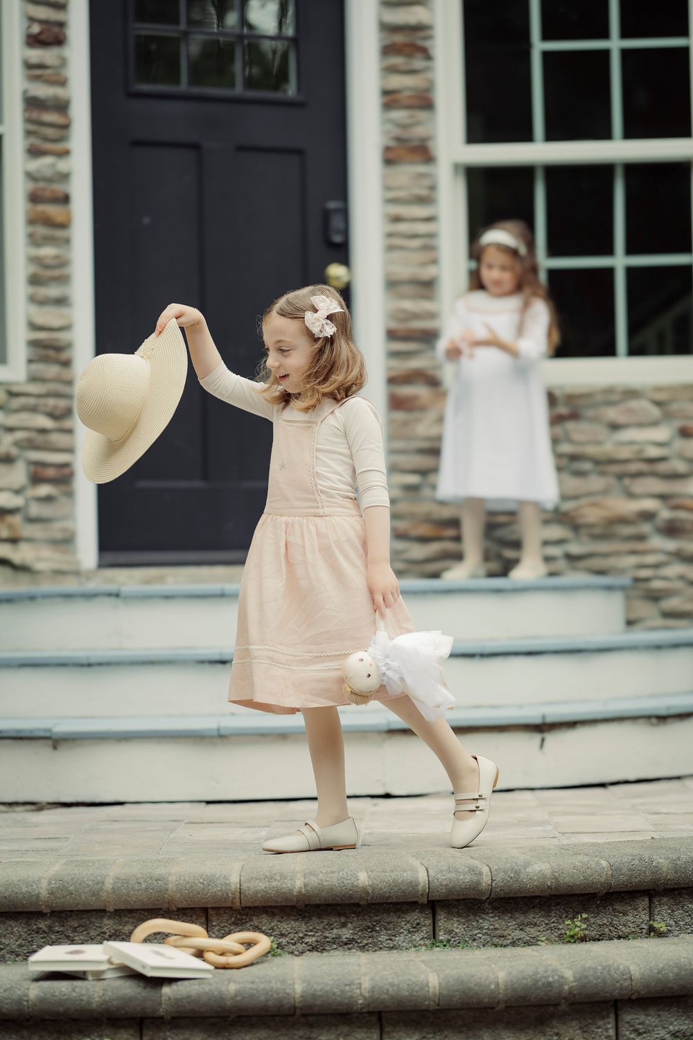 Young girl in pink dress playfully swings straw hat on stone steps while another girl watches from doorway.
