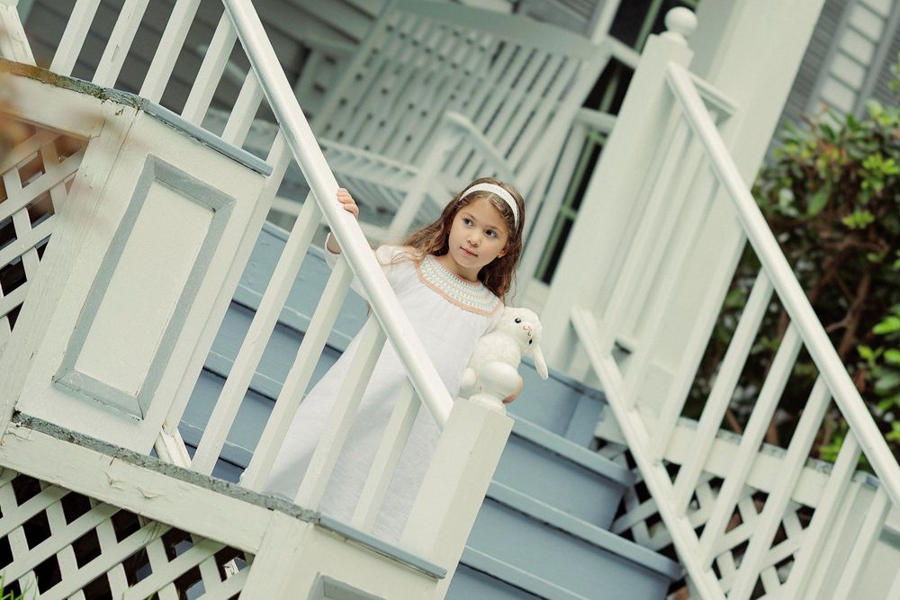 A young girl in a white dress holds a stuffed bunny while standing on blue porch stairs with white railings.