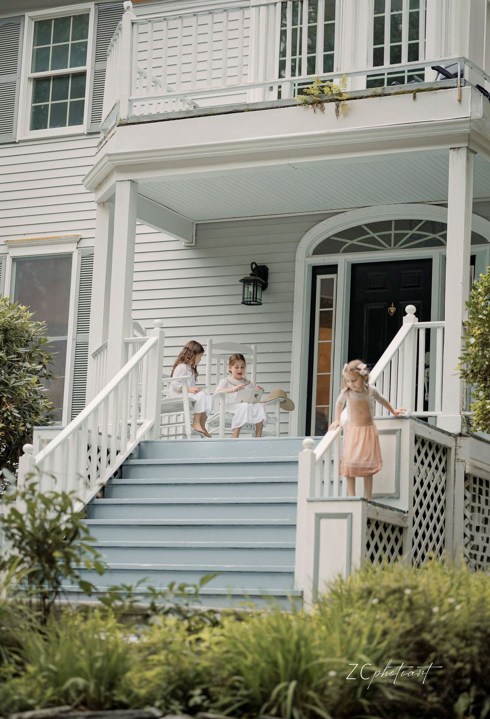 Three young girls in dresses playing on the porch steps of a charming white colonial home.
