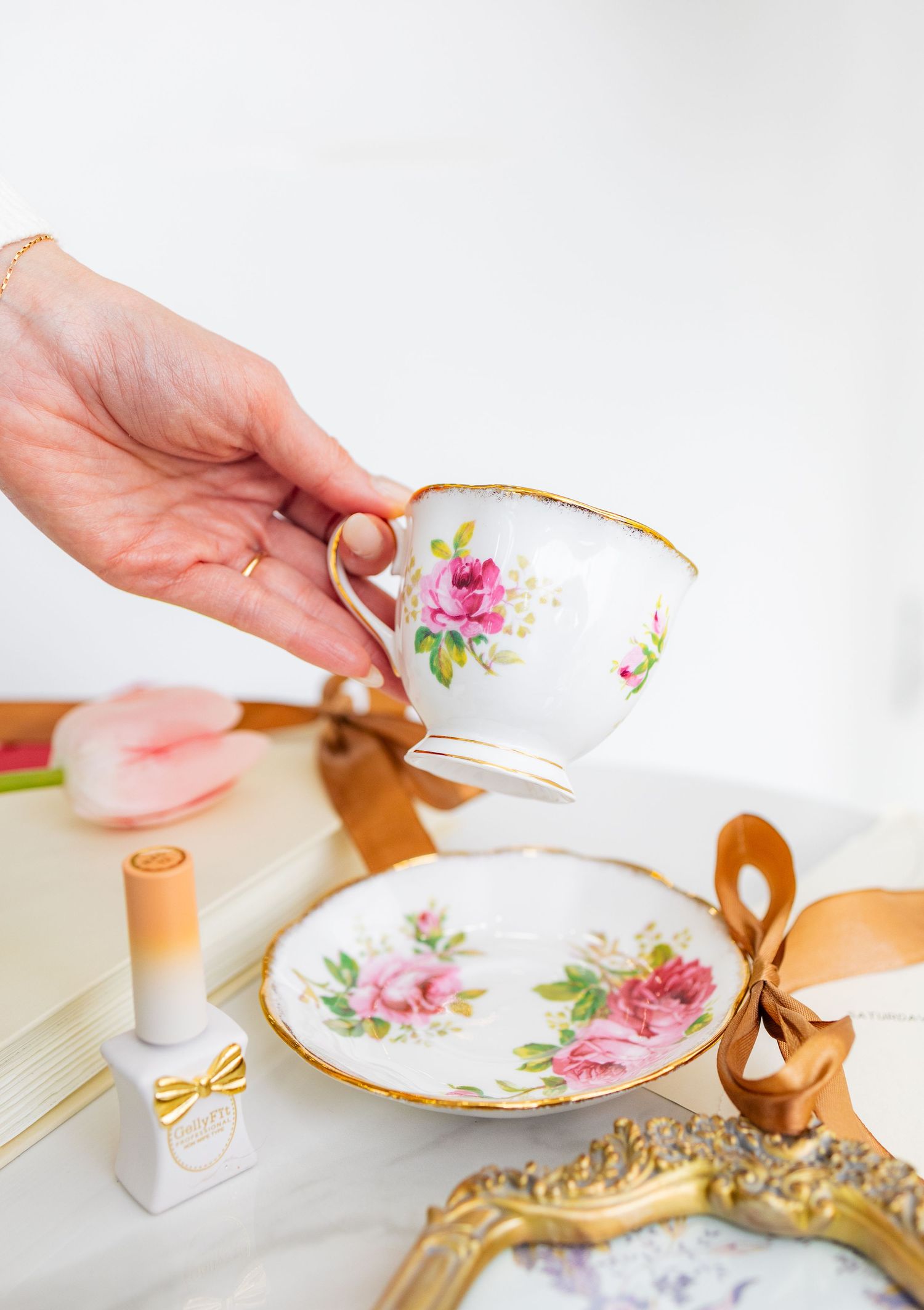 Hand holding a floral porcelain teacup with gold trim above a matching saucer, pink tulip, and ribbon nearby.