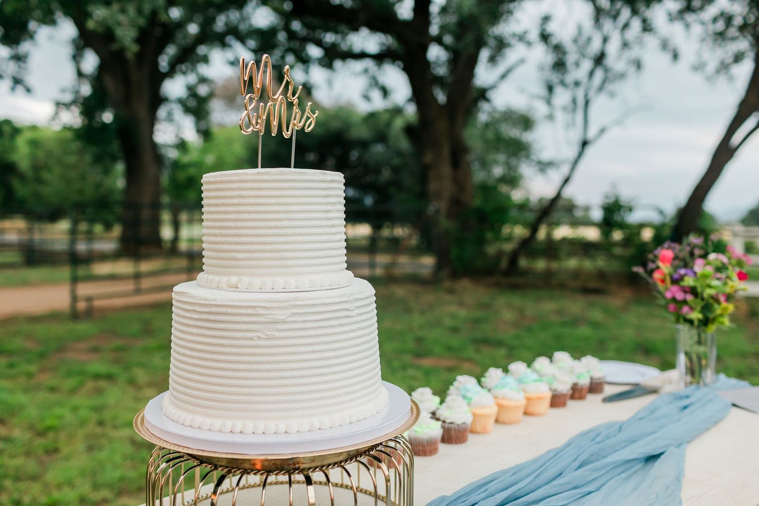 Three-tier white ribbed wedding cake with Mr & Mrs topper displayed outdoors beside pastel cupcakes on a lace table.