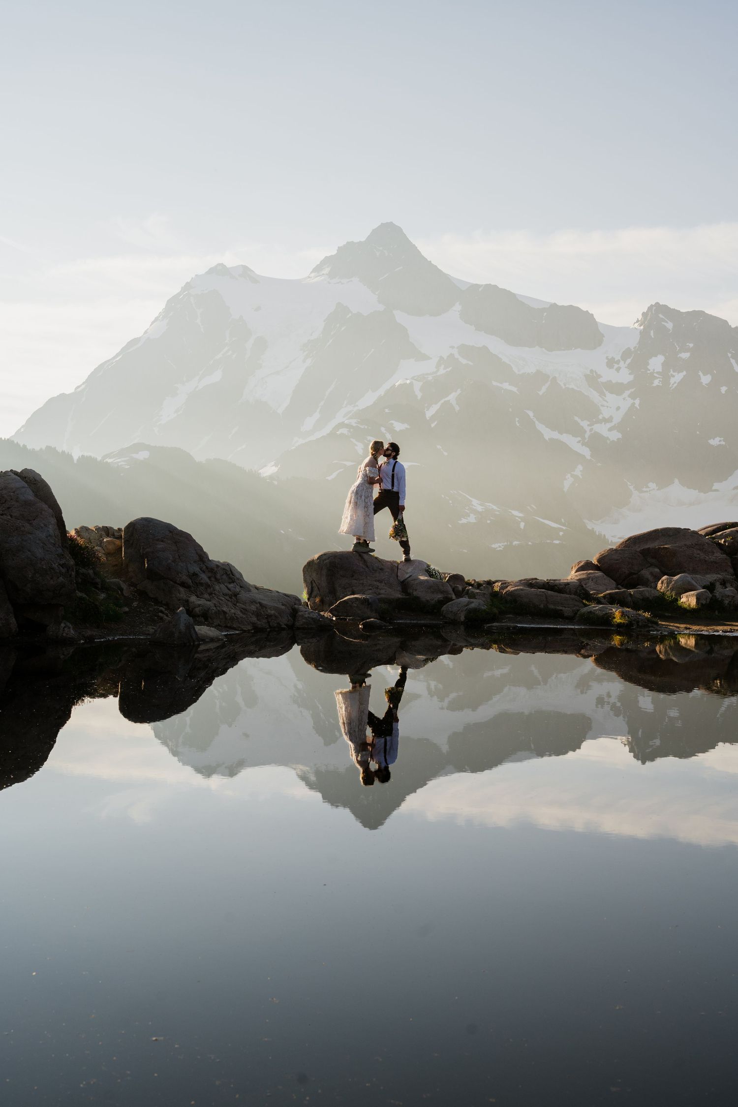 Group celebrating an elopement by a reflective alpine lake with snowy mountain peaks in the background.