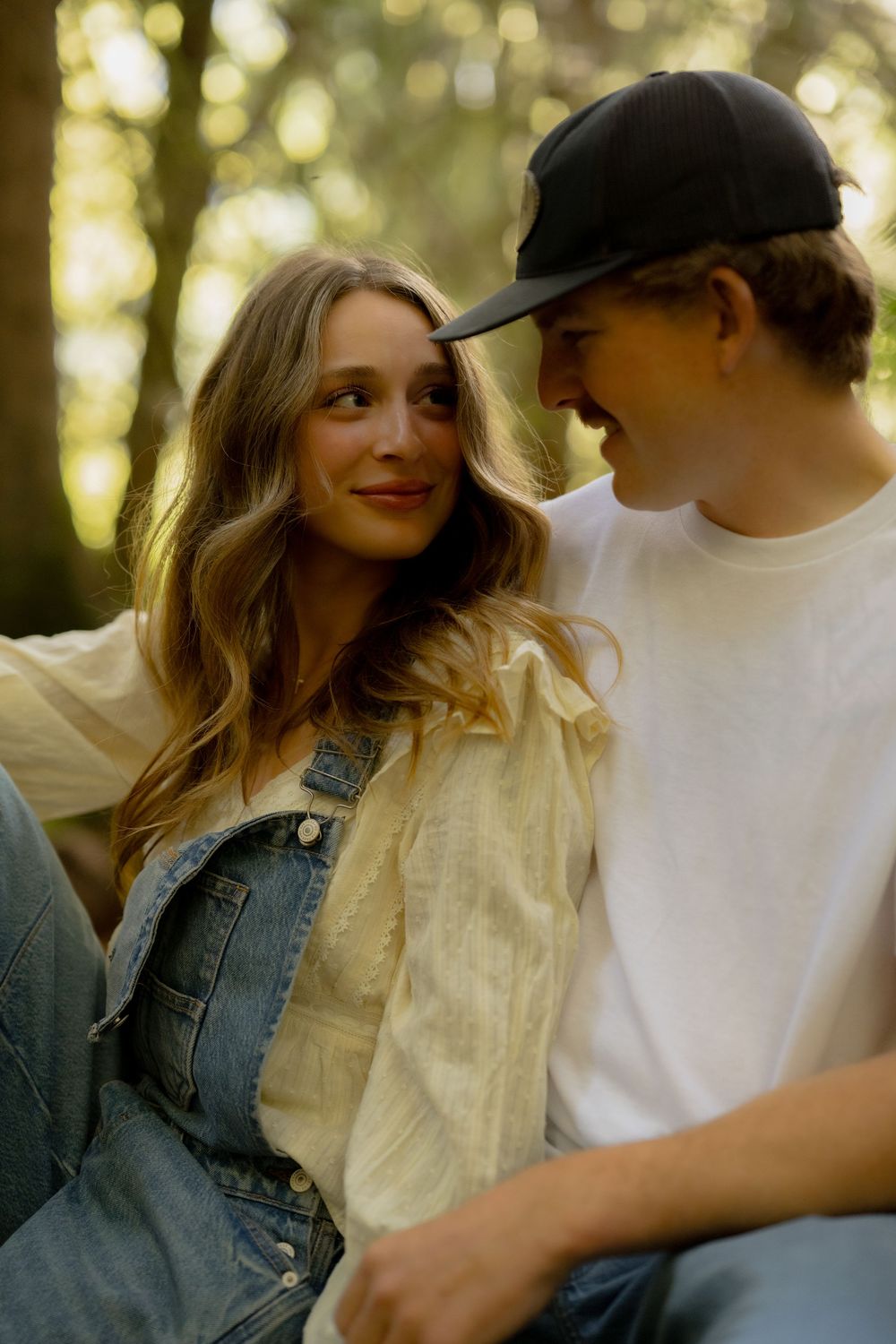 Young couple sitting together in a forest, woman in denim overalls smiling at man in black cap and white tee.