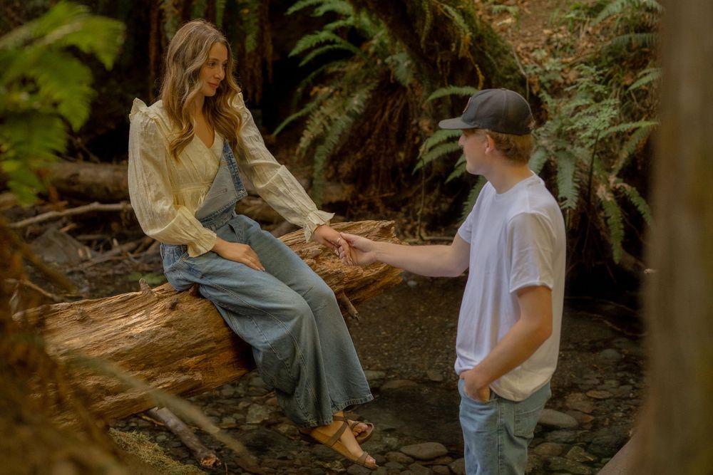 A couple holds hands in a lush forest, woman sitting on a log in a flowy outfit, man in white tee and cap.