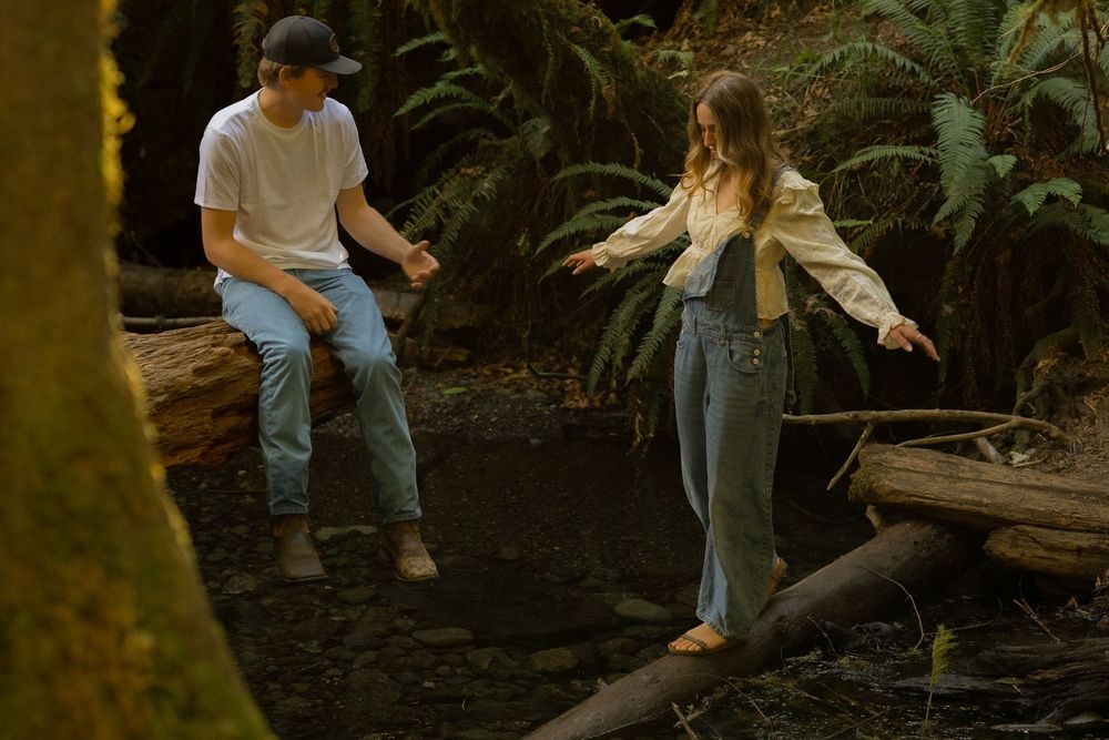 Two young people exploring a lush forest, balancing on fallen logs over a stream among ferns.