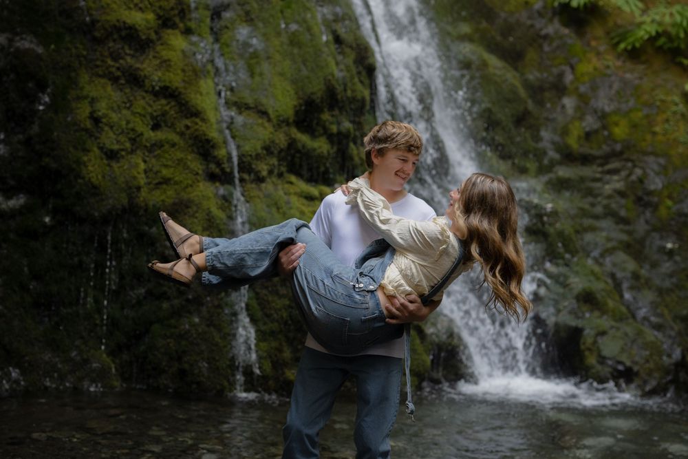 A young couple poses romantically by a mossy waterfall, the man carrying the woman in his arms outdoors.